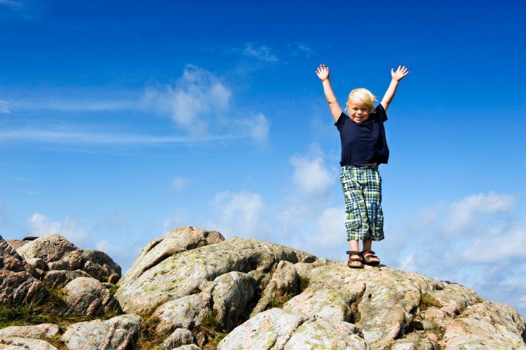 Young boy with his arms raised in victory on top of a rock, conceptual image for conquering challenges, pushing the boundaries, and continuous improvement